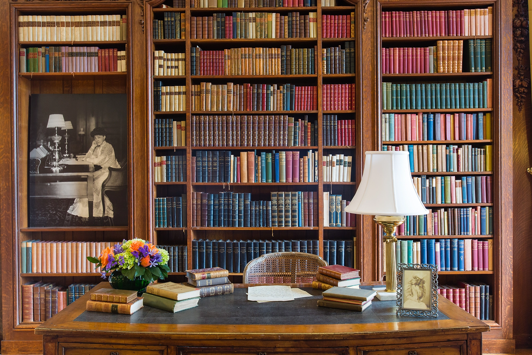 The Library at The Mount, Edith Wharton’s home, featuring floor-to-ceiling wooden bookshelves filled with colorful books. A vintage desk holds stacks of books, a bouquet of flowers, a lamp, and a framed photograph. A black-and-white portrait of Edith Wharton is displayed on the left wall.