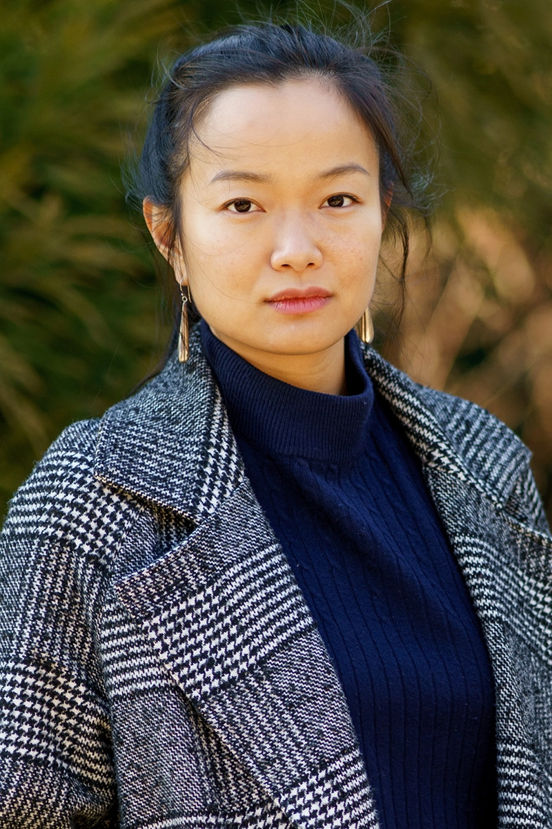 Portrait of a woman, Irene Jiang, with her hair tied back, wearing a houndstooth-patterned coat over a navy blue turtleneck. She gazes confidently at the camera, standing against a blurred natural background.