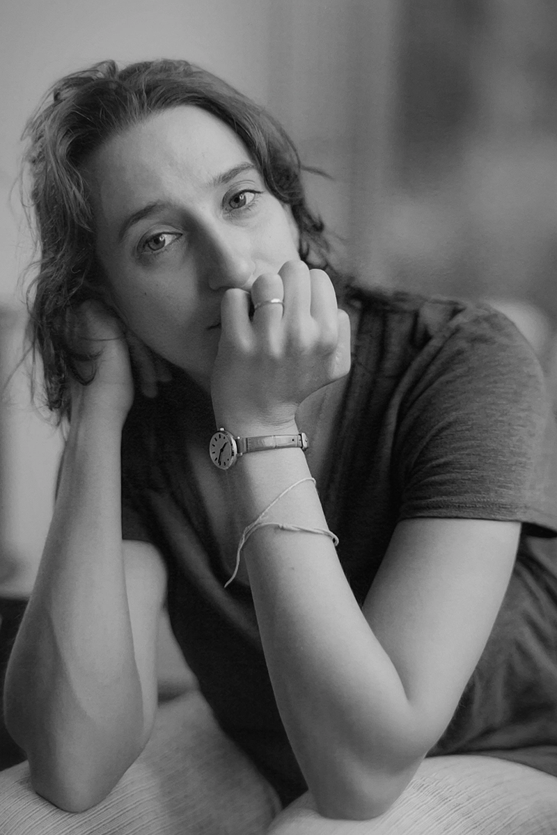 Black-and-white portrait of a woman, Lizzy Beck, resting her chin on her hand. She wears a simple t-shirt, a watch, and a thin bracelet, gazing thoughtfully at the camera against a softly blurred background.