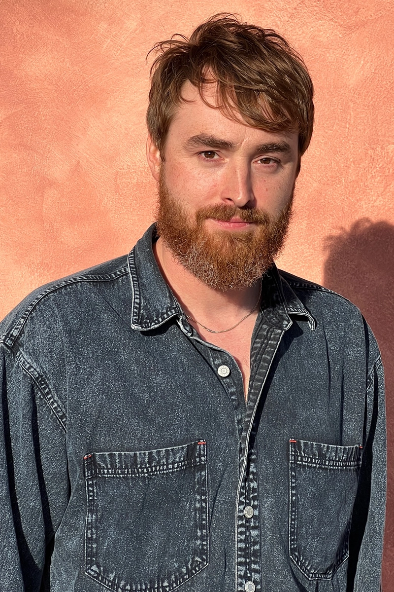 Portrait of a man, Mason Wray, with short brown hair, a reddish beard, and a denim shirt. He stands against a textured peach-colored wall, illuminated by warm sunlight.