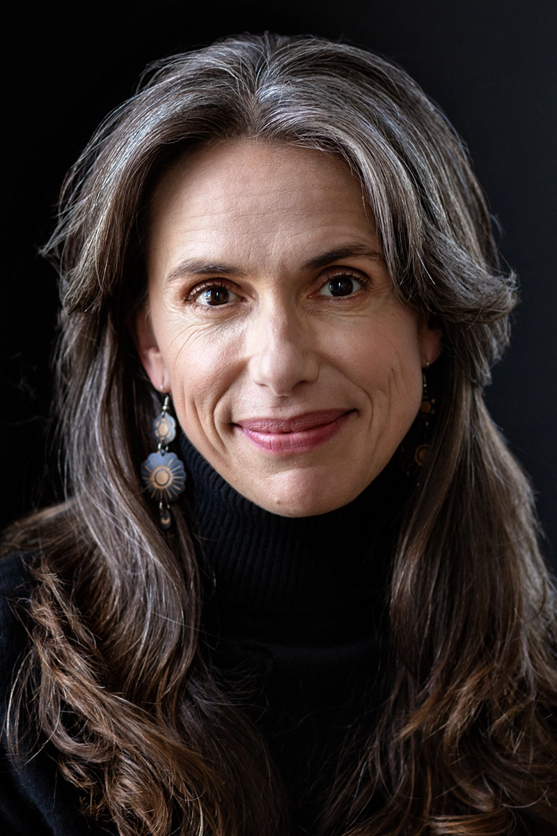 Portrait of a woman, Melenie Freedom-Flynn, with long, wavy hair streaked with silver, wearing a black turtleneck and ornate dangling earrings. She smiles softly against a dark background.