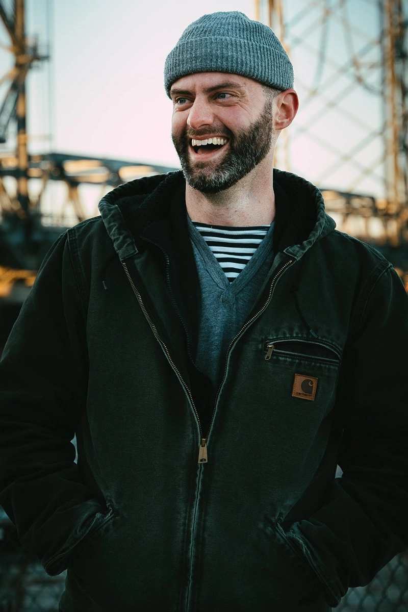 `Portrait of a man, Michael Jerome Plunkett, wearing a gray knit cap, a striped shirt layered under a dark jacket, and smiling broadly. He stands outdoors in front of industrial structures illuminated by soft, natural light.