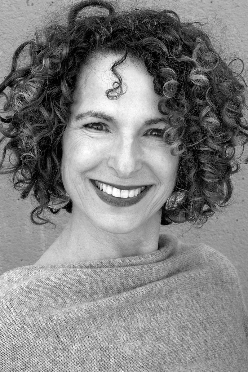 Black-and-white portrait of a smiling woman, Michele Bombardier, with curly hair. She is wearing a light, textured top and is photographed against a plain background.