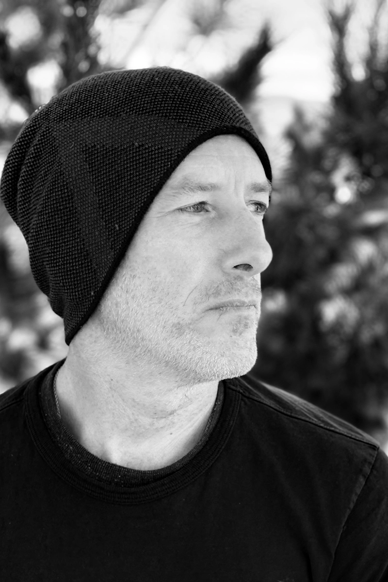 Black-and-white portrait of a man, Thaddeus Haas, wearing a knit beanie and a simple dark shirt. He looks thoughtfully into the distance, with blurred trees in the background.