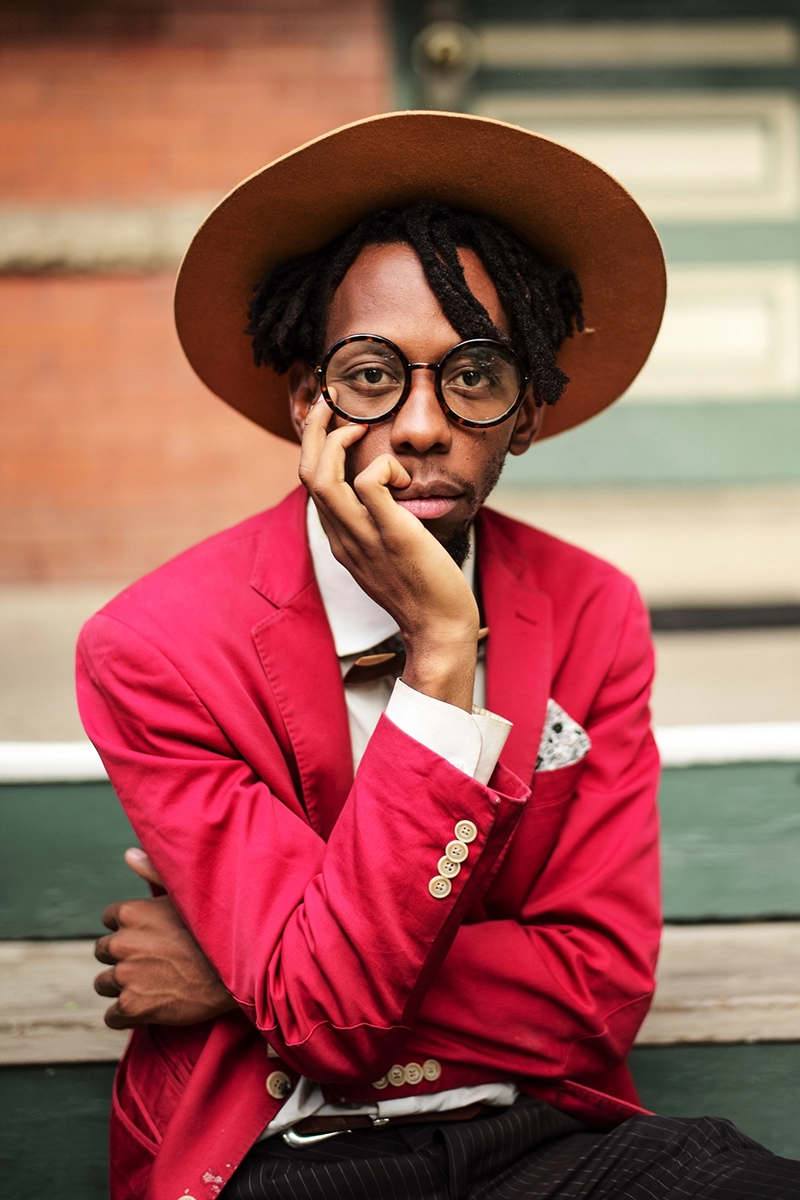 Portrait of a man, Brenton Sizwe Zola, wearing a wide-brimmed hat, round glasses, and a vibrant red blazer over a white shirt with a bow tie. He rests his face on one hand, seated against a blurred background of steps and a brick wall.