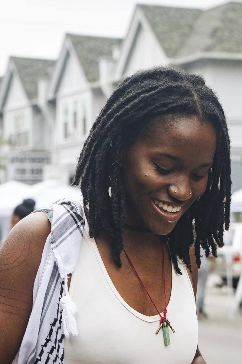 Portrait of a woman, Zite Ezeh, with shoulder-length locs adorned with beads, wearing a white tank top and a green pendant on a red cord. She smiles warmly while standing outdoors against a backdrop of houses and market stalls.