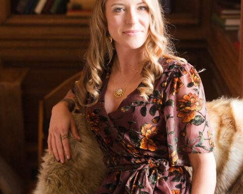 A poised woman with long, wavy blonde hair sits elegantly on a fur-covered chair in a warmly lit library. She wears a flowing, floral-patterned dress in earthy tones, accented by delicate gold jewelry and stacked beaded bracelets. Her gaze is calm and confident, with a subtle smile. Behind her, bookshelves filled with colorful spines add to the cozy, intellectual ambiance. The soft lighting casts a glow, highlighting her serene presence in the space.