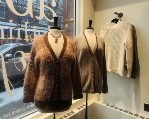 A boutique window display featuring two mannequins dressed in cozy knitwear. The mannequin on the left wears a fuzzy, multicolored brown cardigan with a deep V-neck and dark brown trim, paired with a statement necklace. The mannequin on the right dons a taupe cable-knit cardigan with gold buttons and a long beaded necklace. Behind them, a cream-colored sweater with a textured knit pattern hangs on a wall-mounted rack. The store's large window reflects the snowy street outside, with a parked car and neighboring storefronts visible.