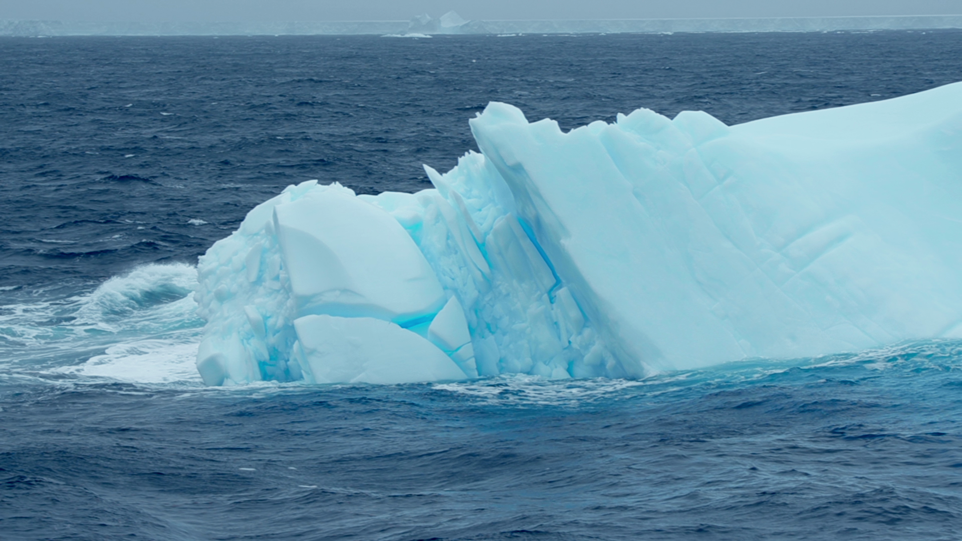 A jagged, vividly blue iceberg floats in the choppy waters of the Southern Ocean, its sharp angles and crevices illuminated by diffuse daylight.