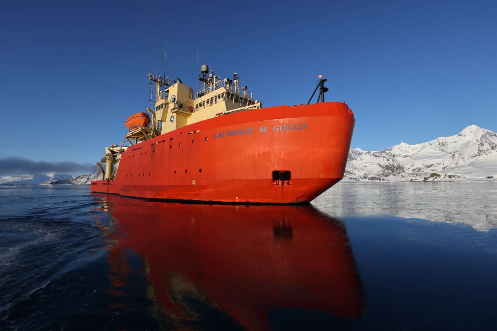 The bright orange hull of the research vessel Laurence M. Gould reflects off the icy surface of the Southern Ocean, with snow-covered mountains in the distance under a clear blue sky.