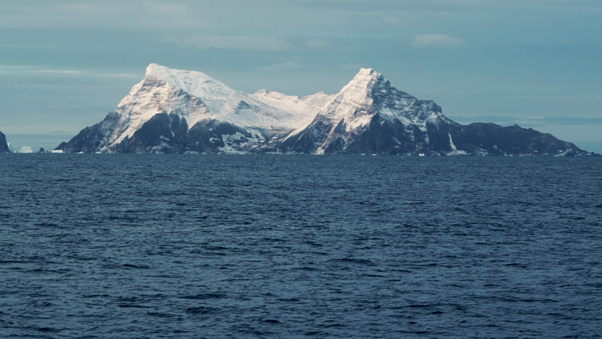A snow-dusted mountain range on South Georgia Island stands against a cloudy sky, viewed across a choppy expanse of dark ocean water.