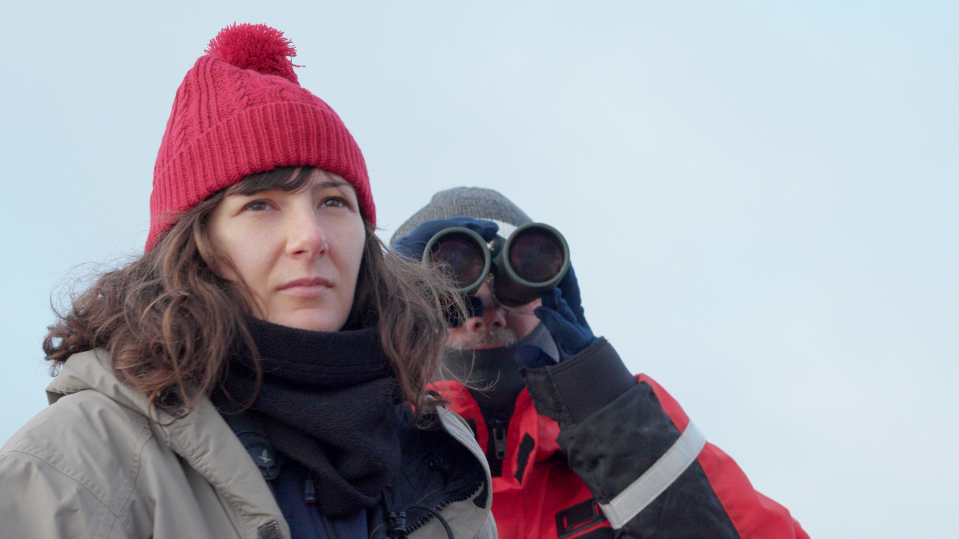 A young woman, Dr. Samantha Monier, wearing a red knit hat and winter gear, looks ahead with a focused expression as fellow researcher Dick Veit peers through binoculars in the background.