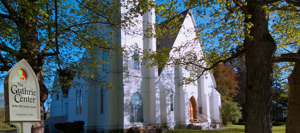 A white wooden church with arched windows and a peaked roof, surrounded by leafy trees under a clear blue sky. A sign in the foreground reads "The Guthrie Center at the Old Trinity Church, Established 1991.