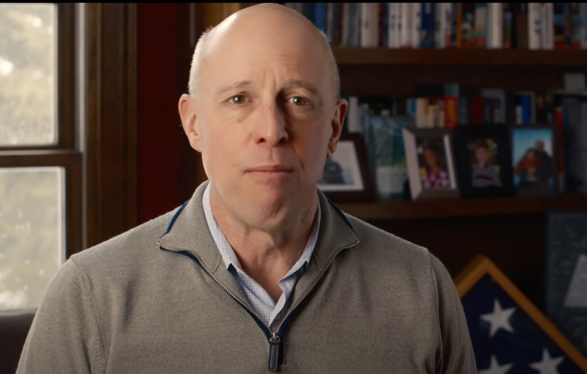 Mike Kennealy, a bald man wearing a light gray quarter-zip sweater over a checkered shirt, appears in a well-lit room with a bookshelf and a folded American flag in the background. Framed family photos and books are visible on the shelf behind him.